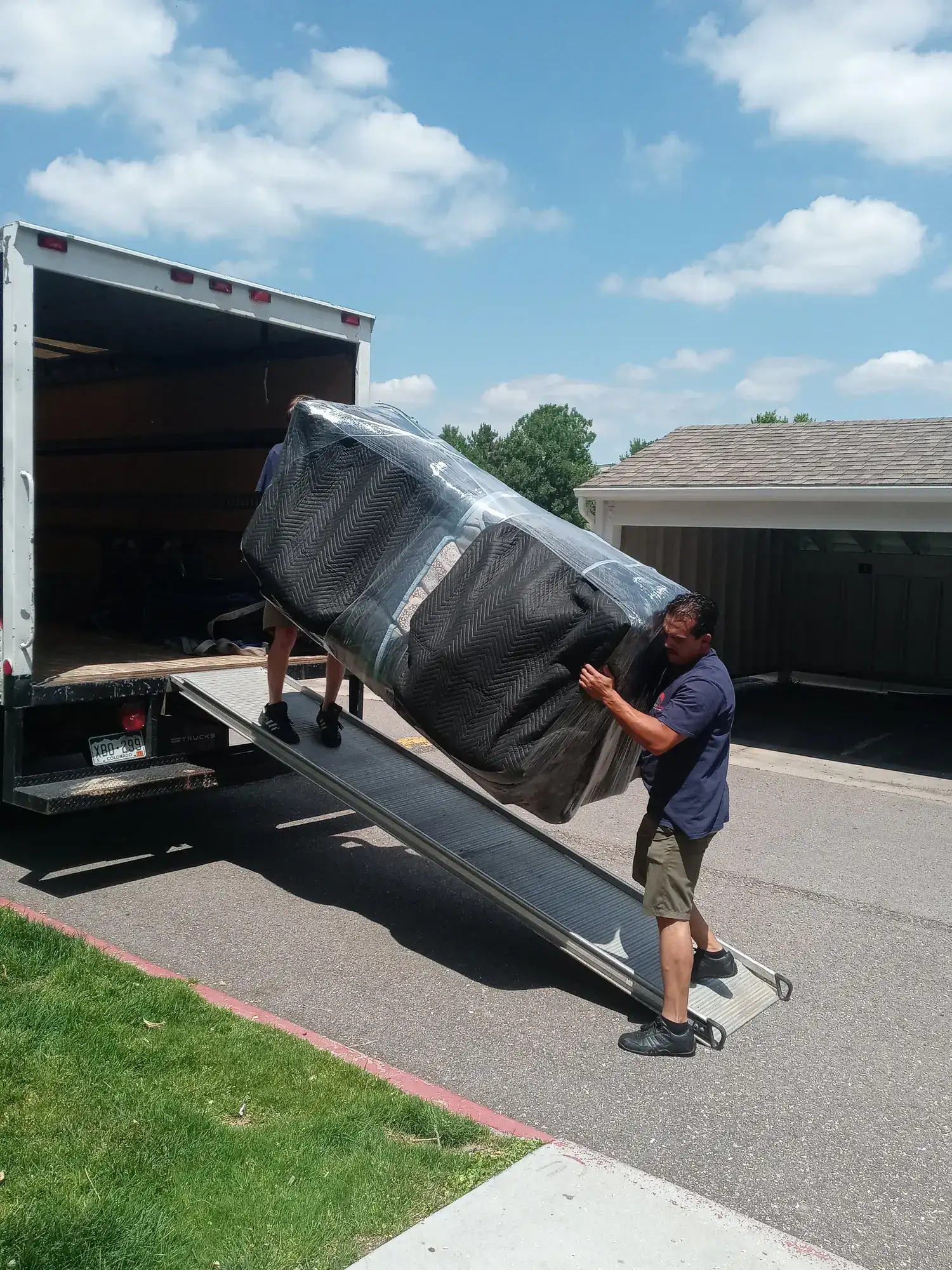 Two movers carrying a shrink-wrapped sofa up a ramp into a moving truck