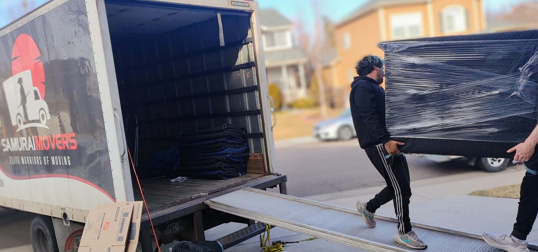 Movers carrying a shrink-wrapped mattress up the ramp into a Samurai Movers truck